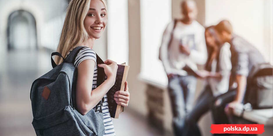 Усміхнена світловолоса дівчина-абітурієнтка з рюкзаком та книжками в руках у коридорі польського навчального закладу - Польська Громада